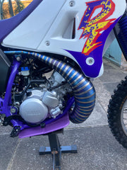 Close-up of a purple and white dirt bike with a branded engine cover on a concrete surface.