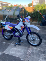 White and purple Yamaha dirt bike on a driveway with a garden and house in the background
